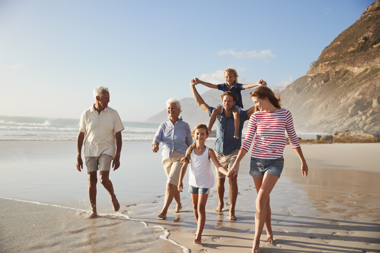 Famille multigénérationnelle marchant ensemble sur une plage, symbole de liens familiaux et de transmission.